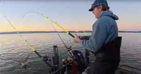 An angler battles a catfish on a neon Hellcat rod at twilight, with the fish’s swirl visible by the boat.
